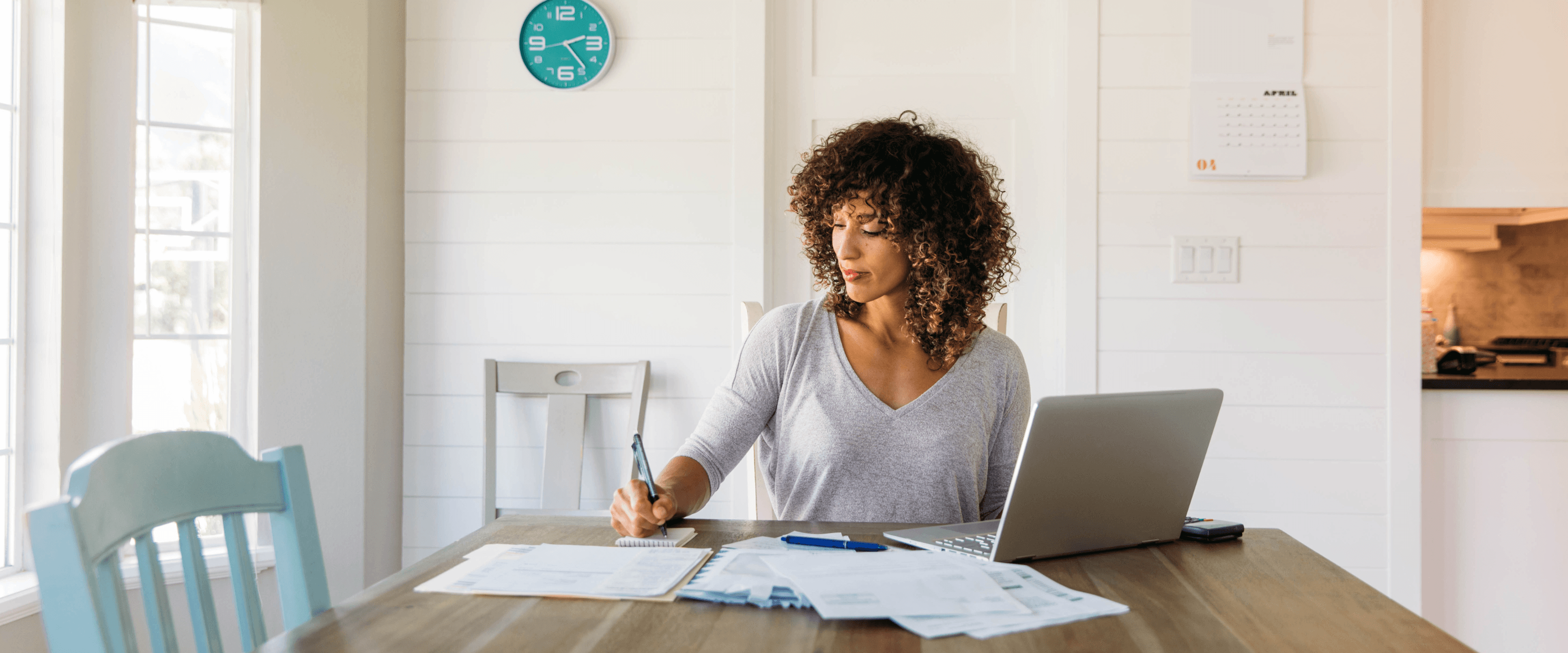 Woman at a dining table reviewing financial paperwork with a laptop and calculator